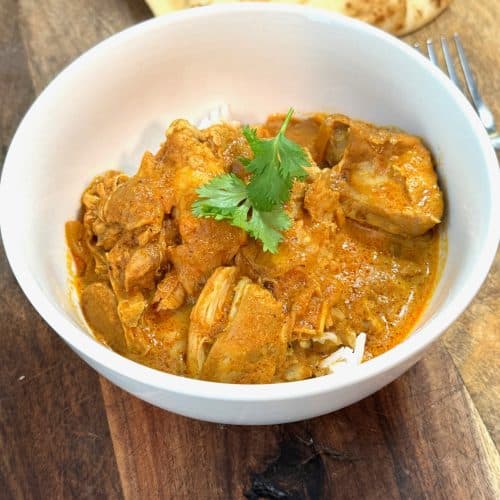 butter chicken in a white bowl sitting on a wooden cutting board next to a piece of naan bread