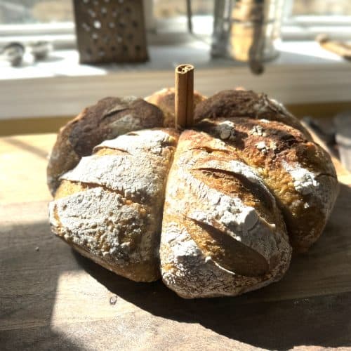 pumpkin shaped sourdough bread on a wooden table in front of a window with a sifter behind it