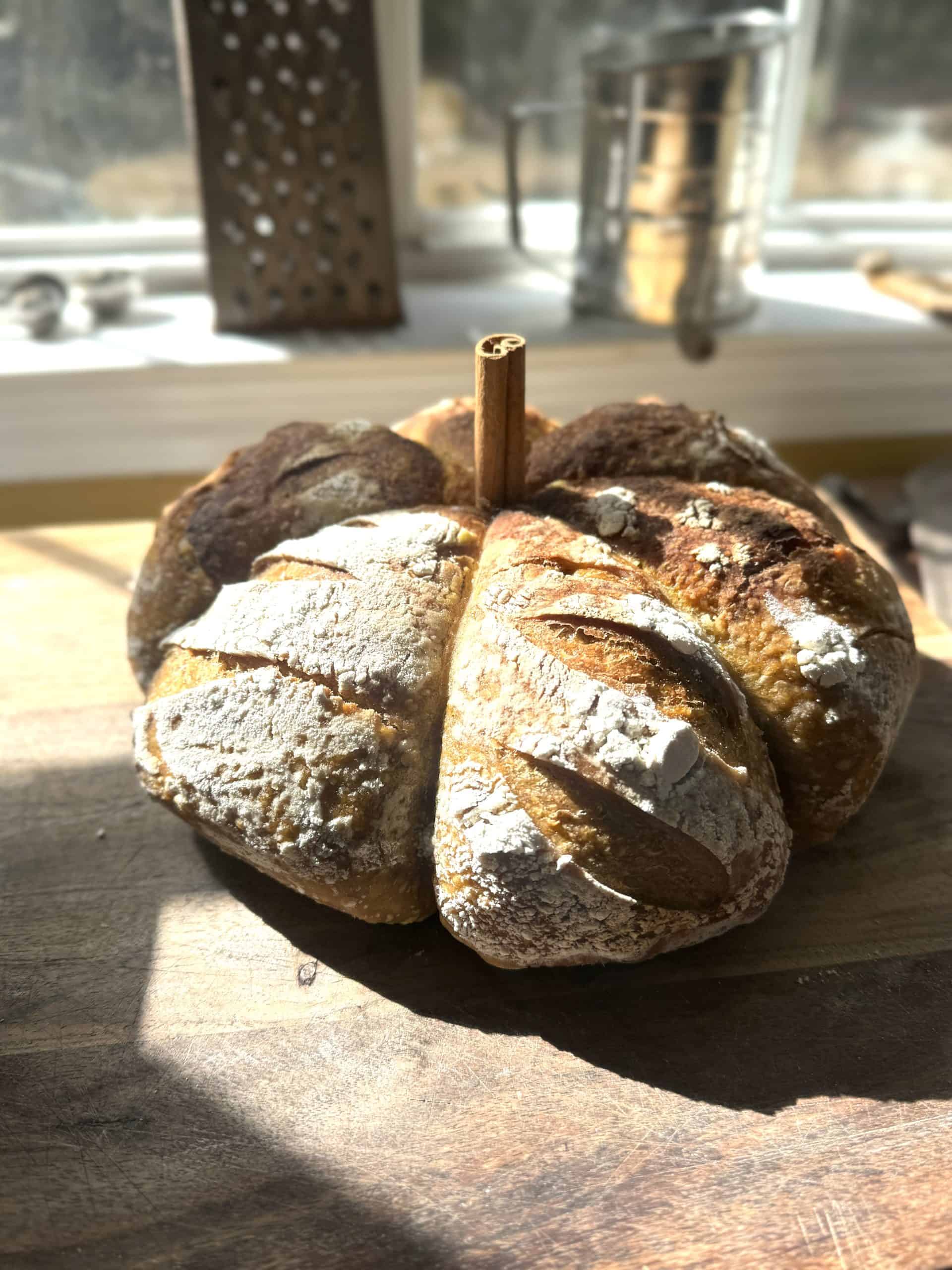 pumpkin shaped sourdough bread on a wooden table in front of a window with a sifter behind it