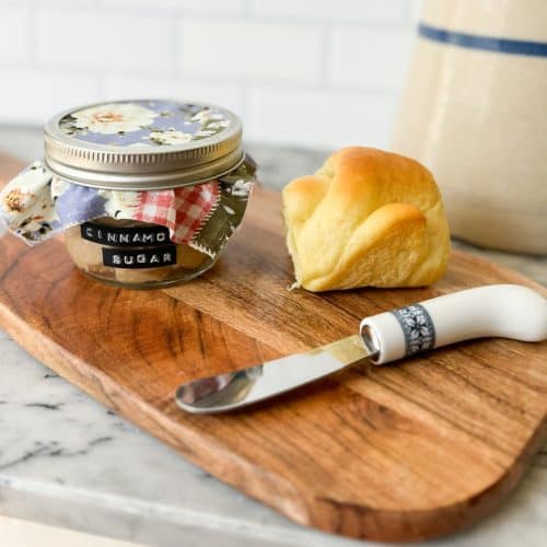 cinnamon sugar butter in a decorative mason jar sitting on a wood cutting board next to a roll and a butter knife