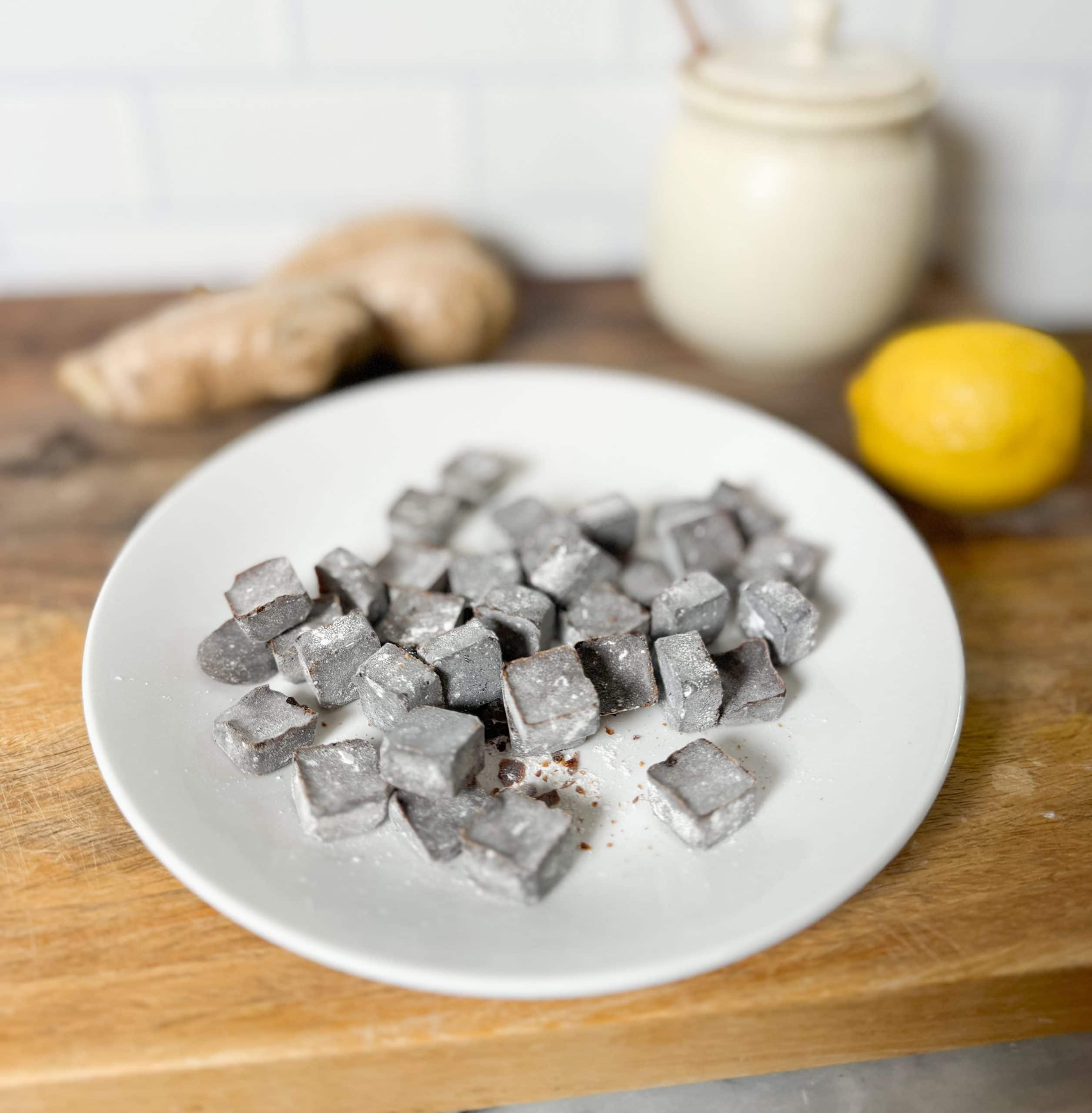 homemade cough drops on a plate sitting on a wooden cutting board with a jar of honey, a lemon and fresh ginger behind it
