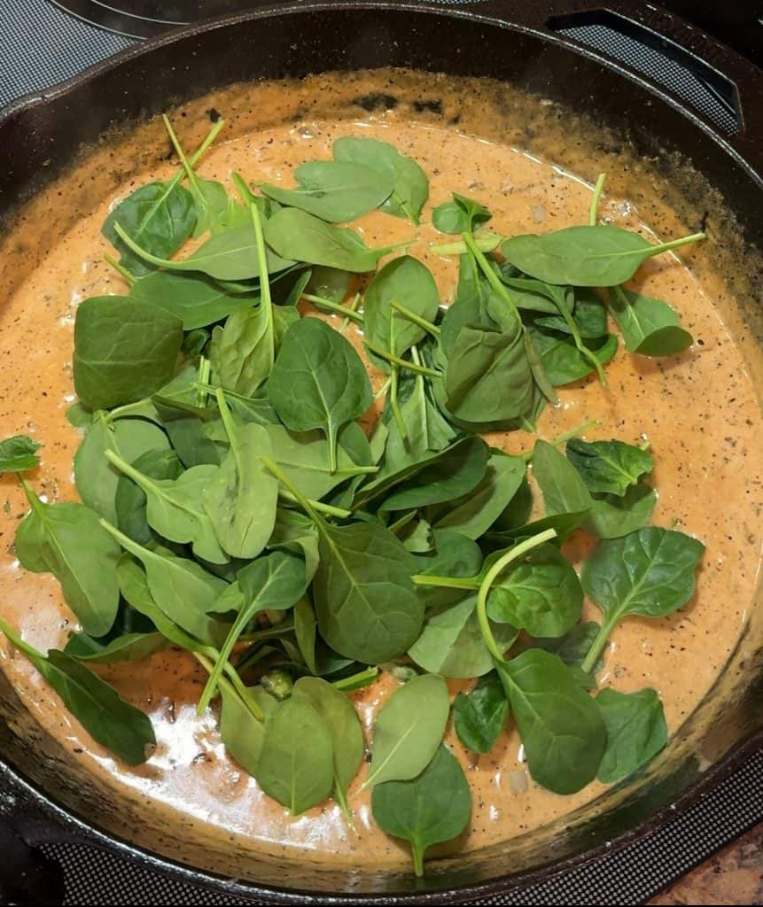 Creamy Tomato Spinach Sauce simmering in a cast iron skillet with fresh spinach being added