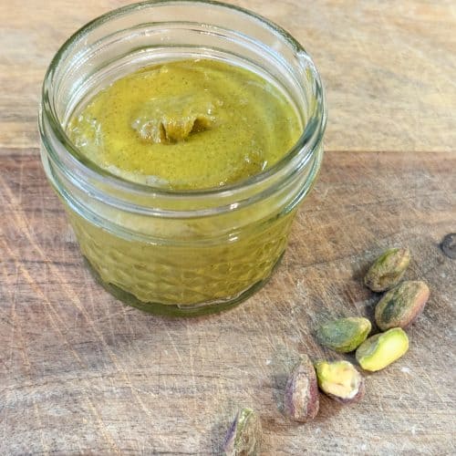 fresh pistachio butter in a small mason jar on a wooden cutting board sitting next to a handful of roasted pistachios