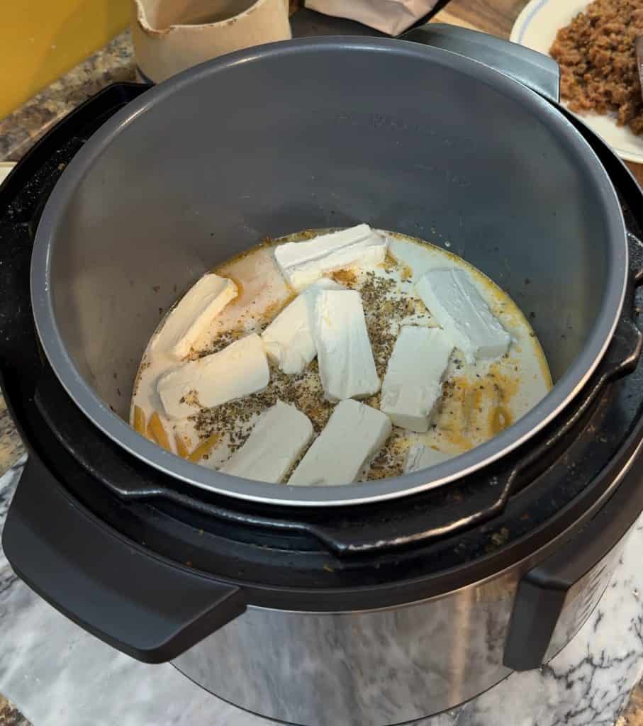 pasta submerged in chicken broth and heavy cream with a layer of sliced cream cheese on top in an instant pot