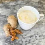 Homemade Golden Milk in a white mug sitting next to a turmeric root and a ginger root