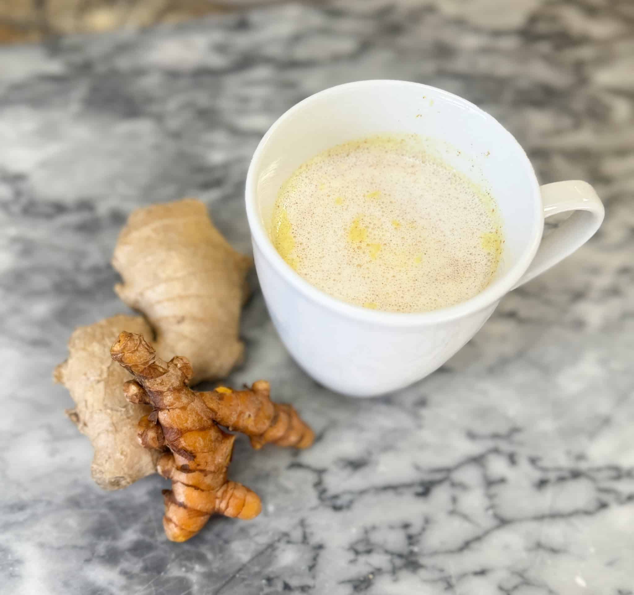 Homemade Golden Milk in a white mug sitting next to a turmeric root and a ginger root