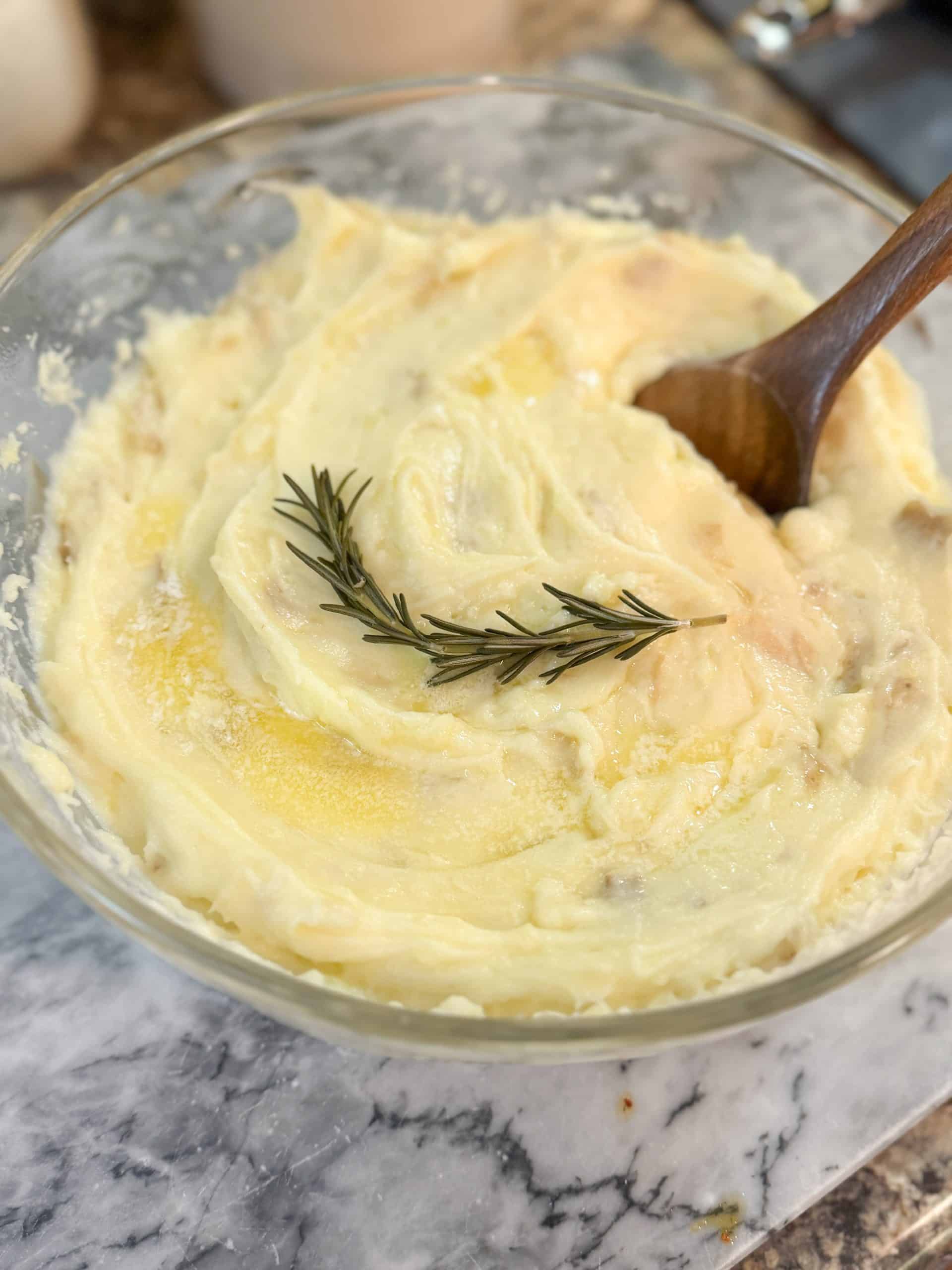Instant Pot Creamy Garlic Mashed Potatoes in a large glass bowl with a wooden spoon and a sprig of rosemary