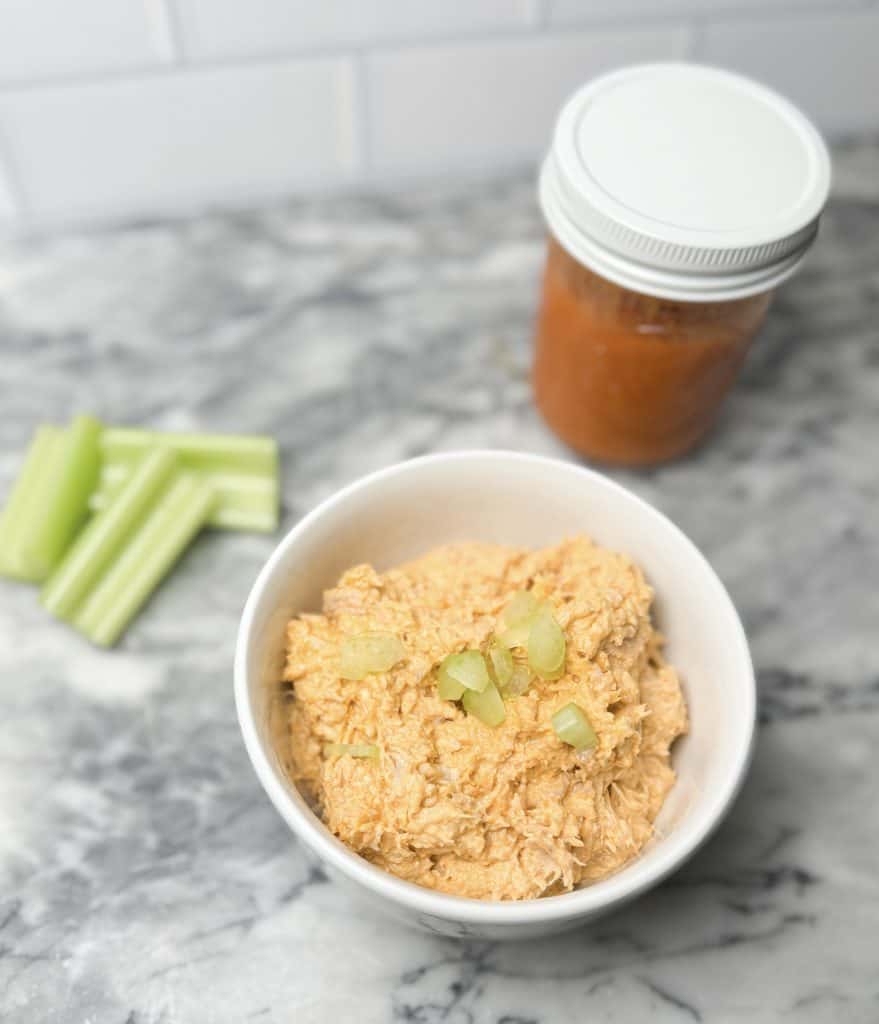hot honey chicken salad in a bowl on a marble counter sitting next to a jar of buffalo sauce and some celery