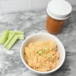 hot honey chicken salad in a bowl on a marble counter sitting next to a jar of buffalo sauce and some celery