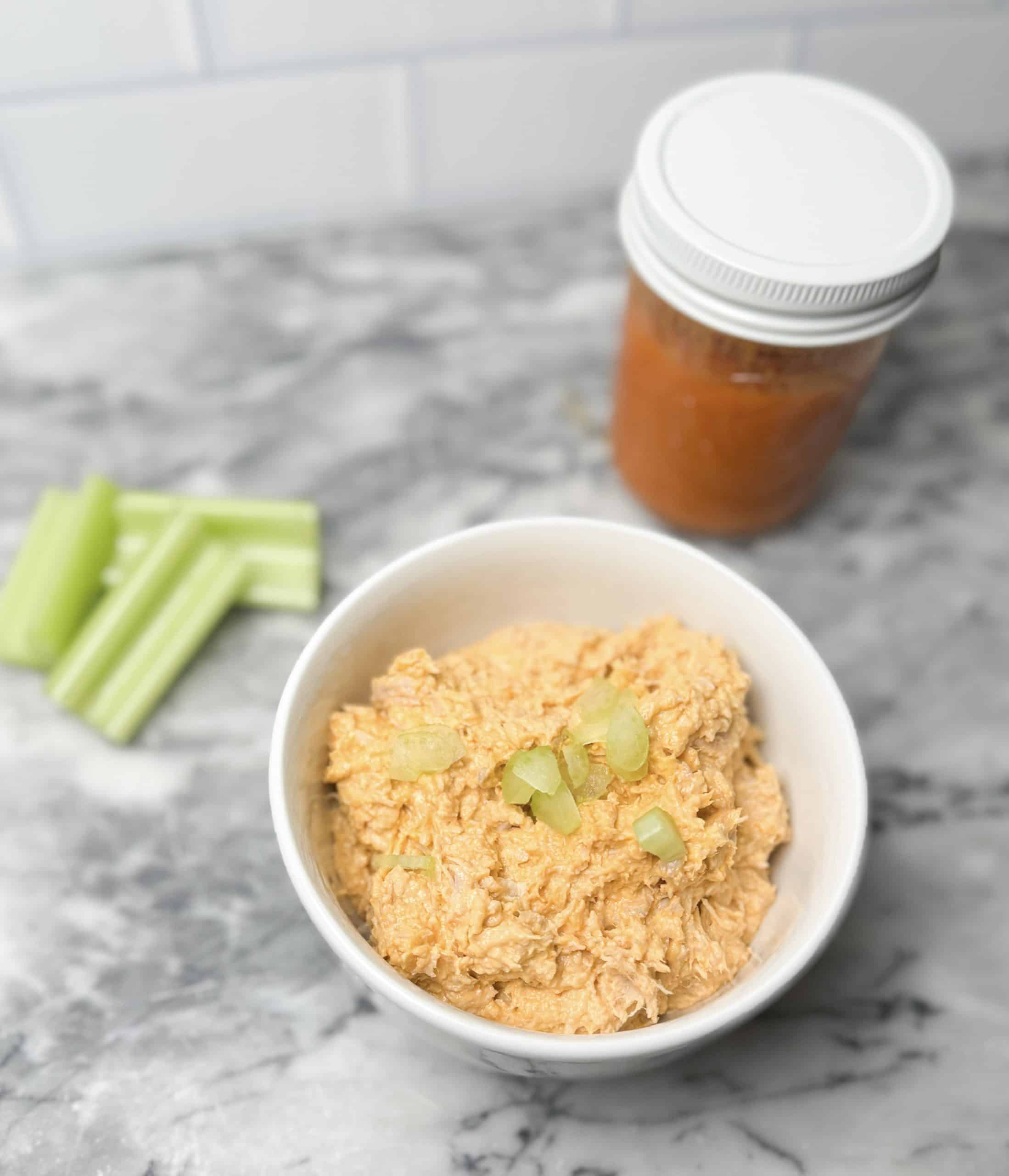 hot honey chicken salad in a bowl on a marble counter sitting next to a jar of buffalo sauce and some celery