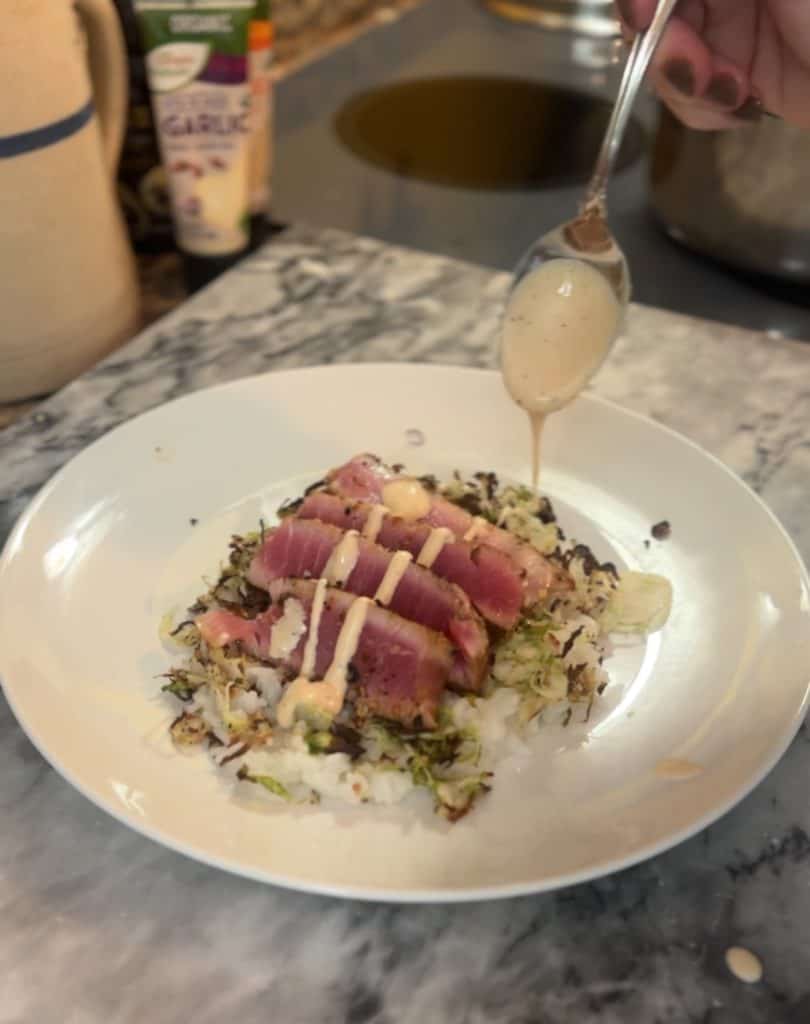 drizzling a creamy sauce over sliced ahi tuna steaks sitting on shredded brussels sprouts on a bed of rice