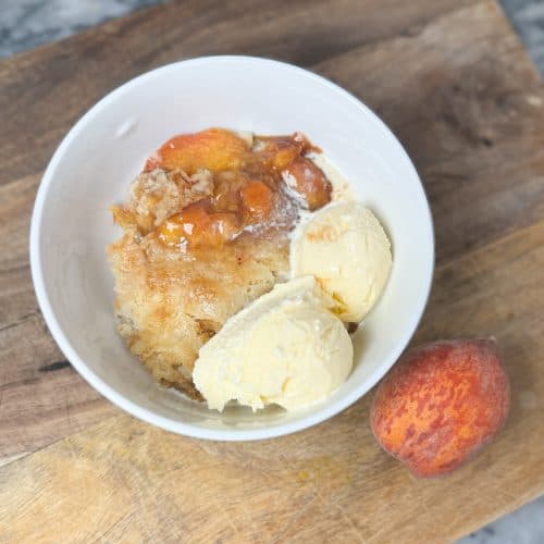 instant pot peach cobbler in a white bowl with 2 scoops of vanilla ice cream sitting next to a whole peach