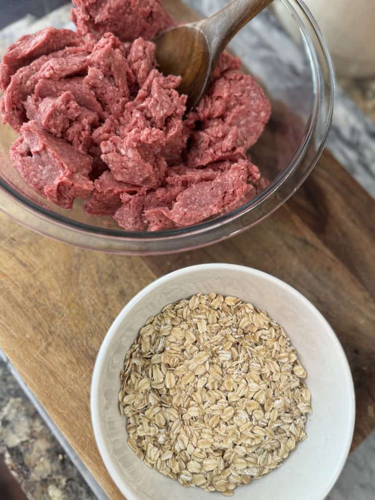 Hamburger meat in a large bowl sitting next to a smaller bowl of old fashioned oats